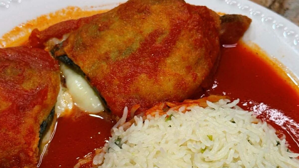 A close-up of a traditional Mexican chile relleno oozing with melted mozzarella cheese, covered in a red guajillo and tomato broth, served alongside white rice on a white plate.